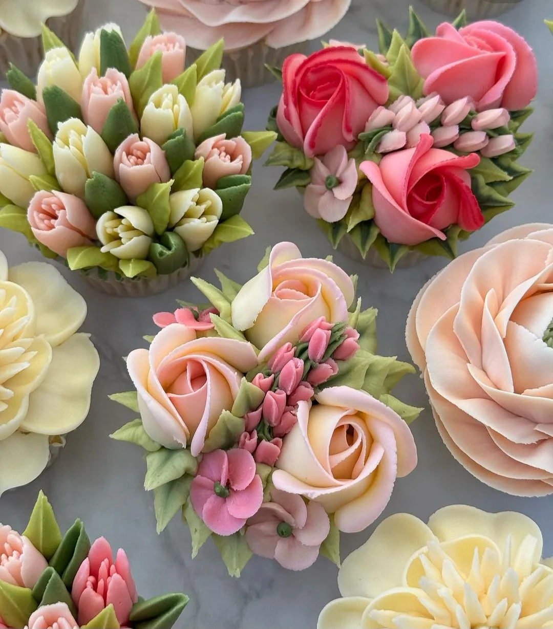 Piped buttercream flower cupcakes in pink, cream and green, including tulips, roses and ranunculus, arranged on marble.