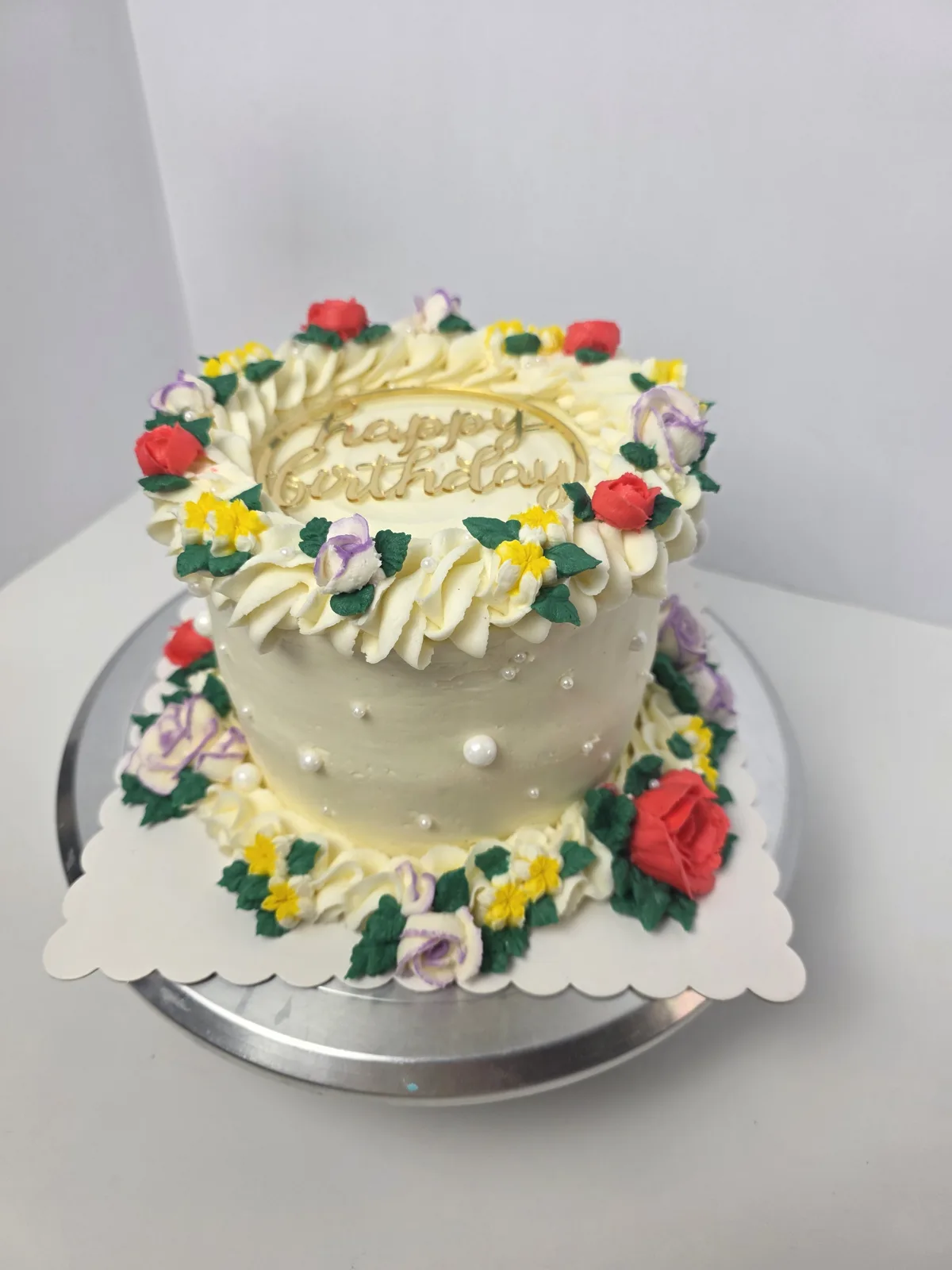 Cream frosted round cake topped with a buttercream wreath of red roses, lavender roses, yellow blooms, and green leaves, with gold "happy birthday" lettering and white pearl accents.