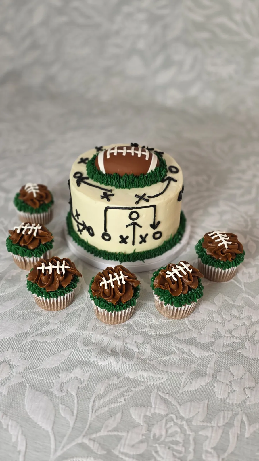 Small white frosted cake decorated with black football play diagrams and topped with a fondant football on green piped grass, surrounded by matching cupcakes.