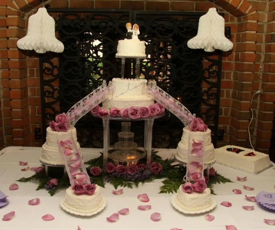Multi tiered white wedding cake with purple rose decorations, lace ribbon bridges, bride and groom topper, and a small fountain centerpiece.