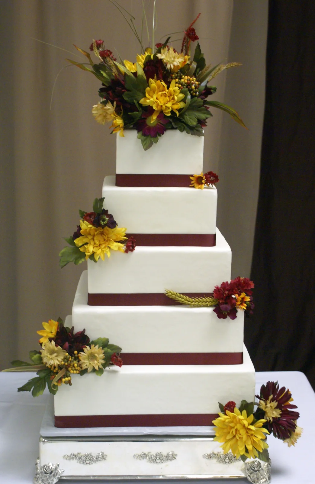Four tiered square white cake wrapped with burgundy ribbons and decorated with cascading yellow and burgundy floral arrangements and greenery.