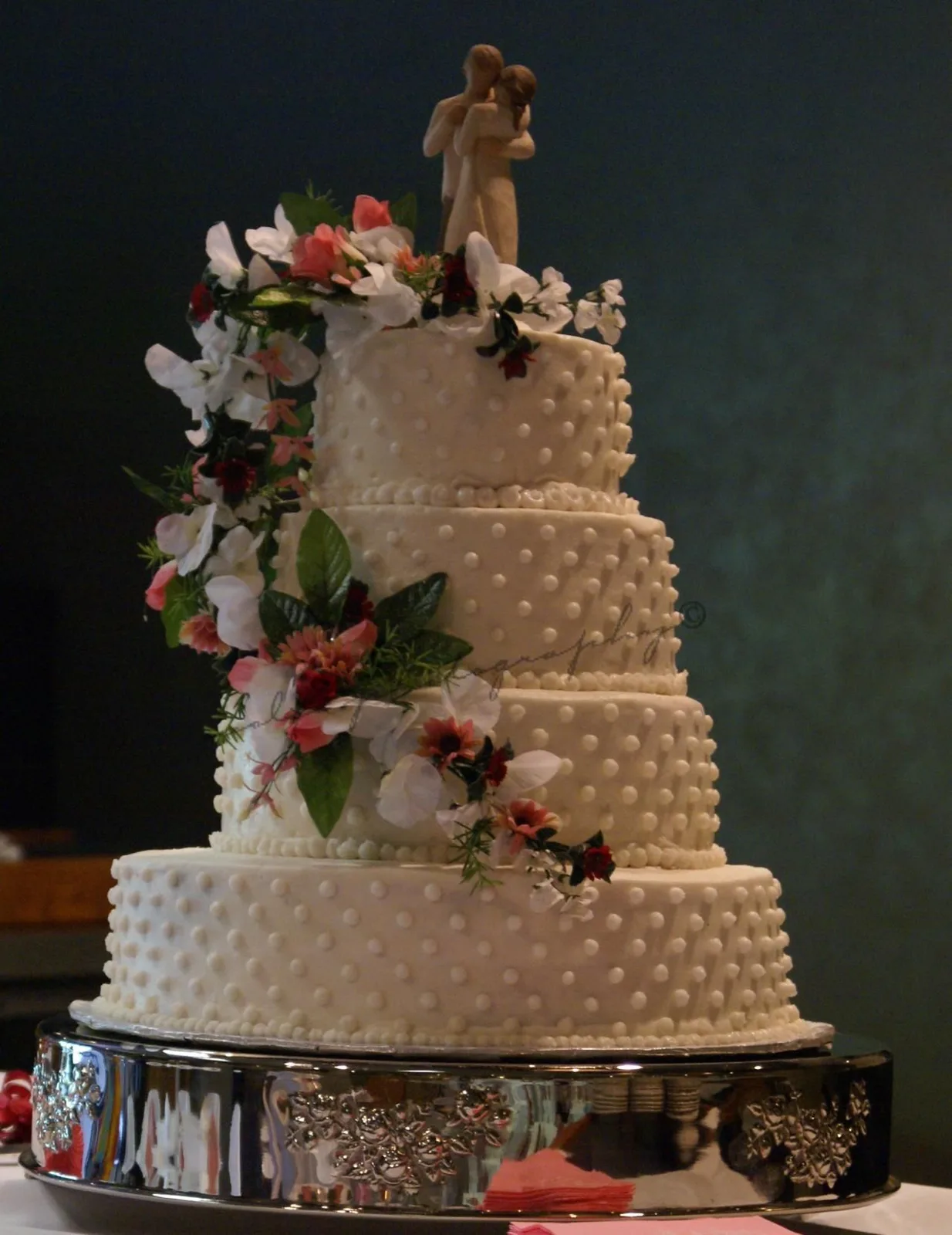 Four tier white wedding cake with raised polka dot texture, cascading white, pink, and red sugar flowers with greenery, topped with a bride and groom figurine.
