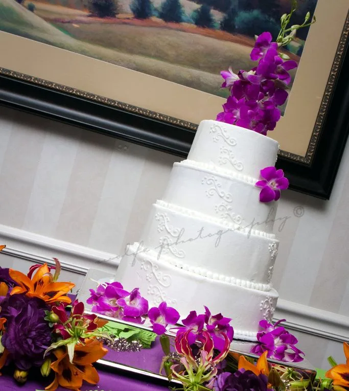 A four tiered white wedding cake decorated with delicate piped scrollwork, accented by tall purple orchids and a vibrant arrangement of orange and purple blooms at the base.