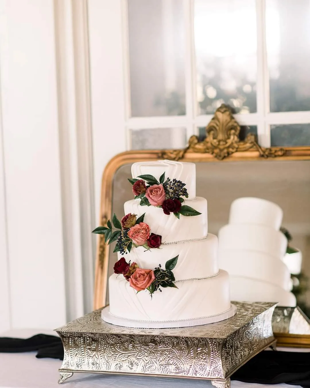A four tiered white cake with vertical textured lines features arrangements of pink roses, dark red flowers, and green leaves. It rests on a silver embossed stand in front of a gold framed mirror.