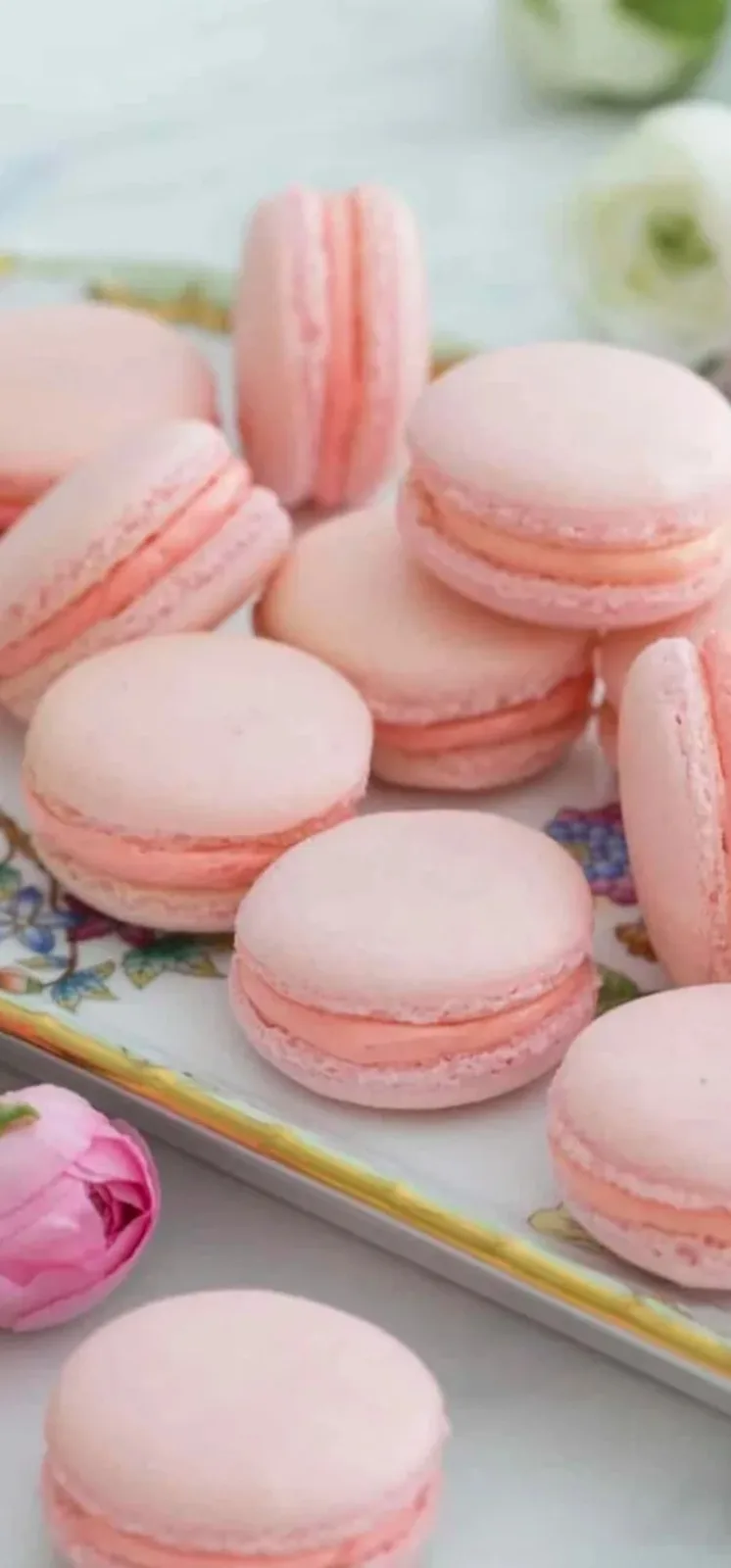 Pink macarons with coral filling stacked on a floral-patterned plate, surrounded by white and pink flowers.