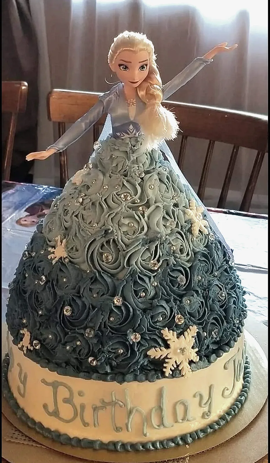 Tall dome cake with blue rosette frosting, silver gem accents, white snowflake decorations, and a blonde princess figurine wearing a blue bodice on top.