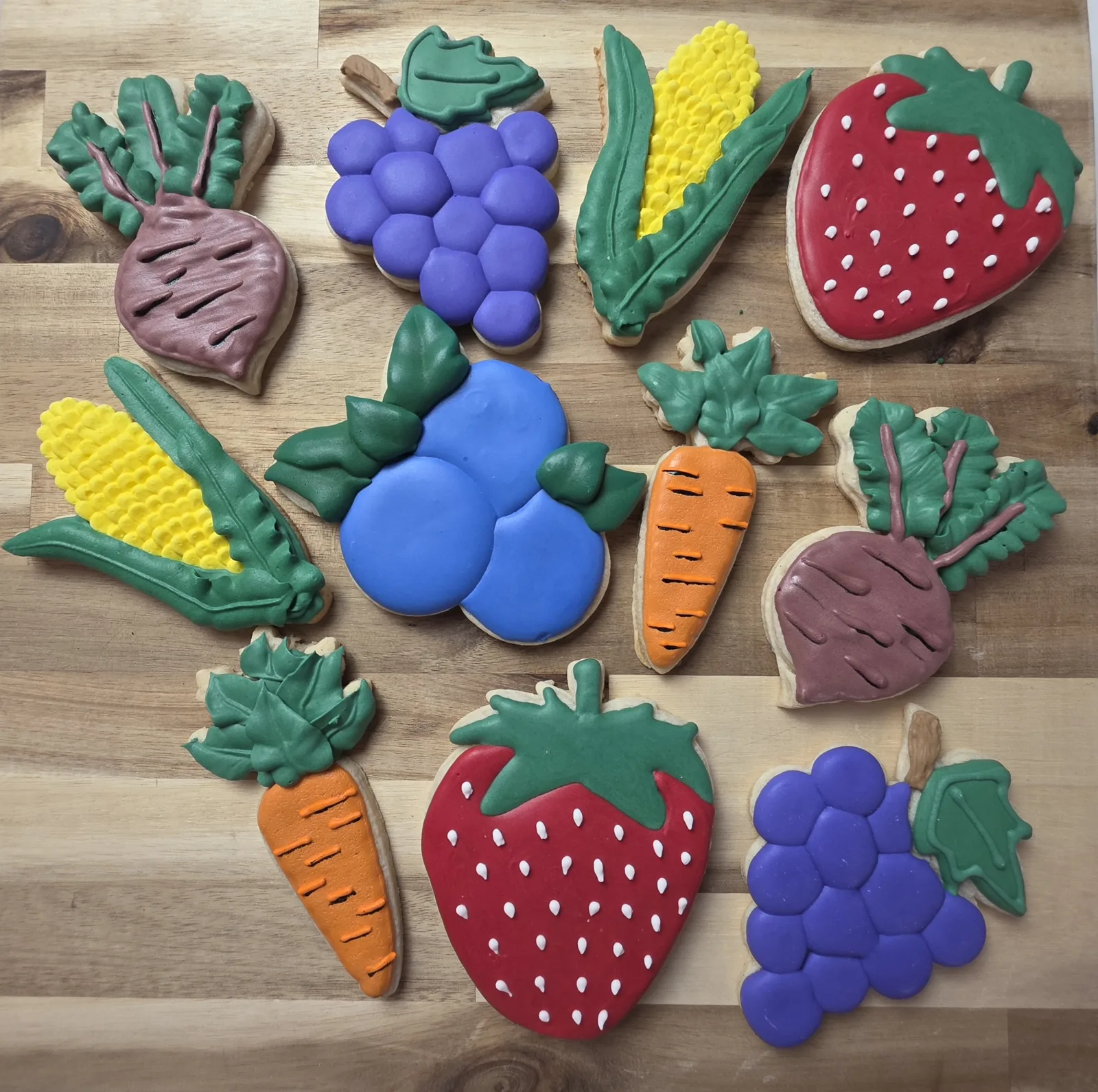 Assorted sugar cookies shaped like strawberries, corn, beets, grapes, blueberries, and carrots, decorated with bright red, yellow, purple, blue, orange, and green icing on a wooden surface.
