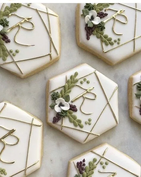 White hexagon cookies with a gold monogram R, greenery and white flowers on marble.