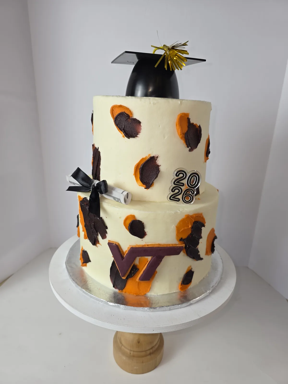 Two tier white frosted cake with orange and maroon paint splatters, topped with a black graduation cap and gold tassel, decorated with a rolled diploma, "2026" emblem, and maroon VT logo.