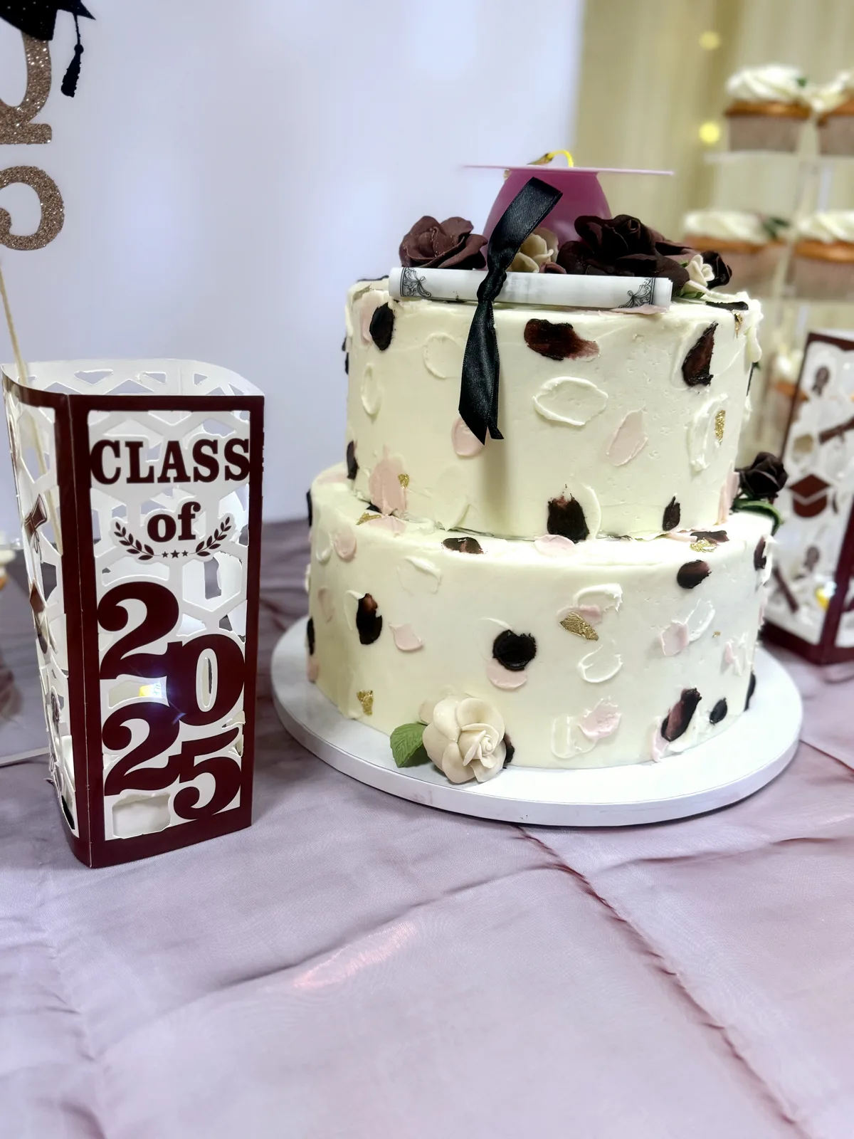 Two tier white buttercream cake with burgundy and blush brushstroke details, gold leaf accents, topped with a pink graduation cap, burgundy roses, and a rolled diploma.