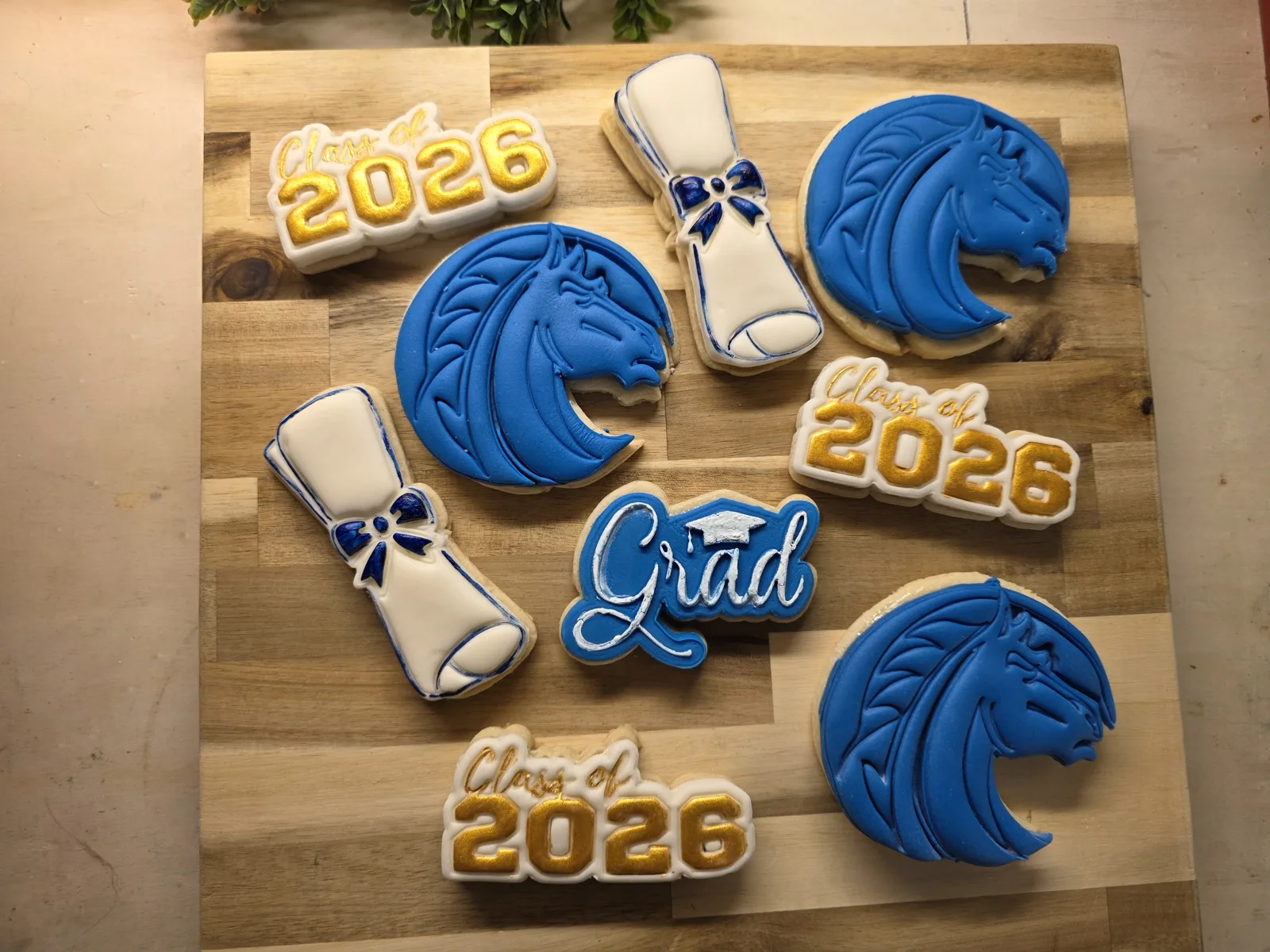 Decorated sugar cookies featuring blue dragon designs, white diploma scrolls with blue bows, gold Class of 2026 plaques, and a blue Grad sign on a wooden board.