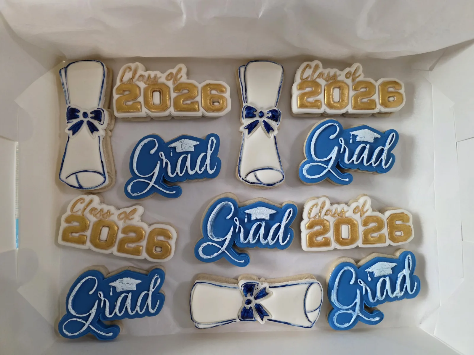 Boxed set of graduation sugar cookies iced in navy, gold and white, reading Grad, Class of 2026, with diploma scroll shapes.