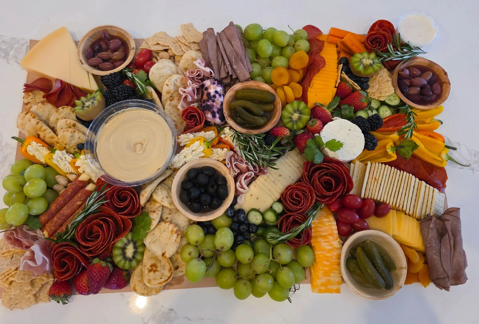 Charcuterie board with cheeses, salami roses, crackers, grapes, strawberries, kiwi, olives, pickles, and hummus.
