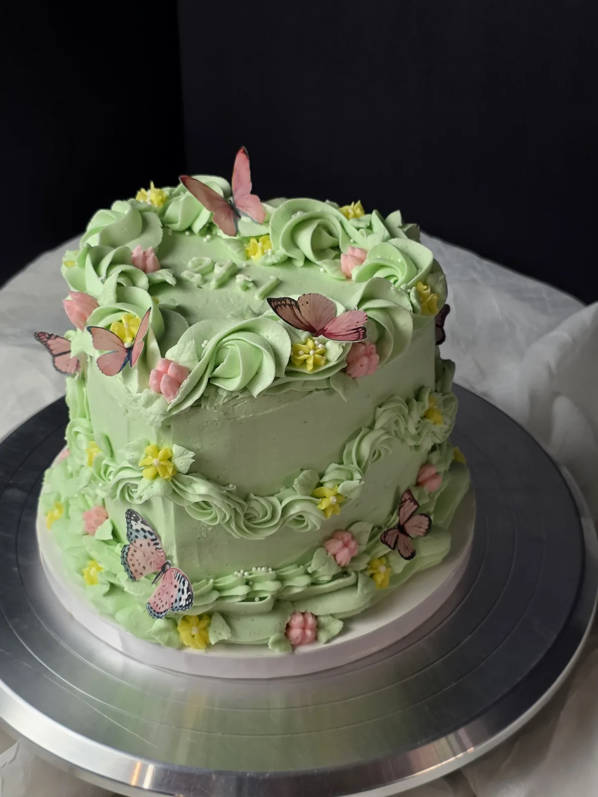 Pale green frosted round cake decorated with piped buttercream roses, small yellow and pink blossoms, and pink paper butterflies on a silver board.