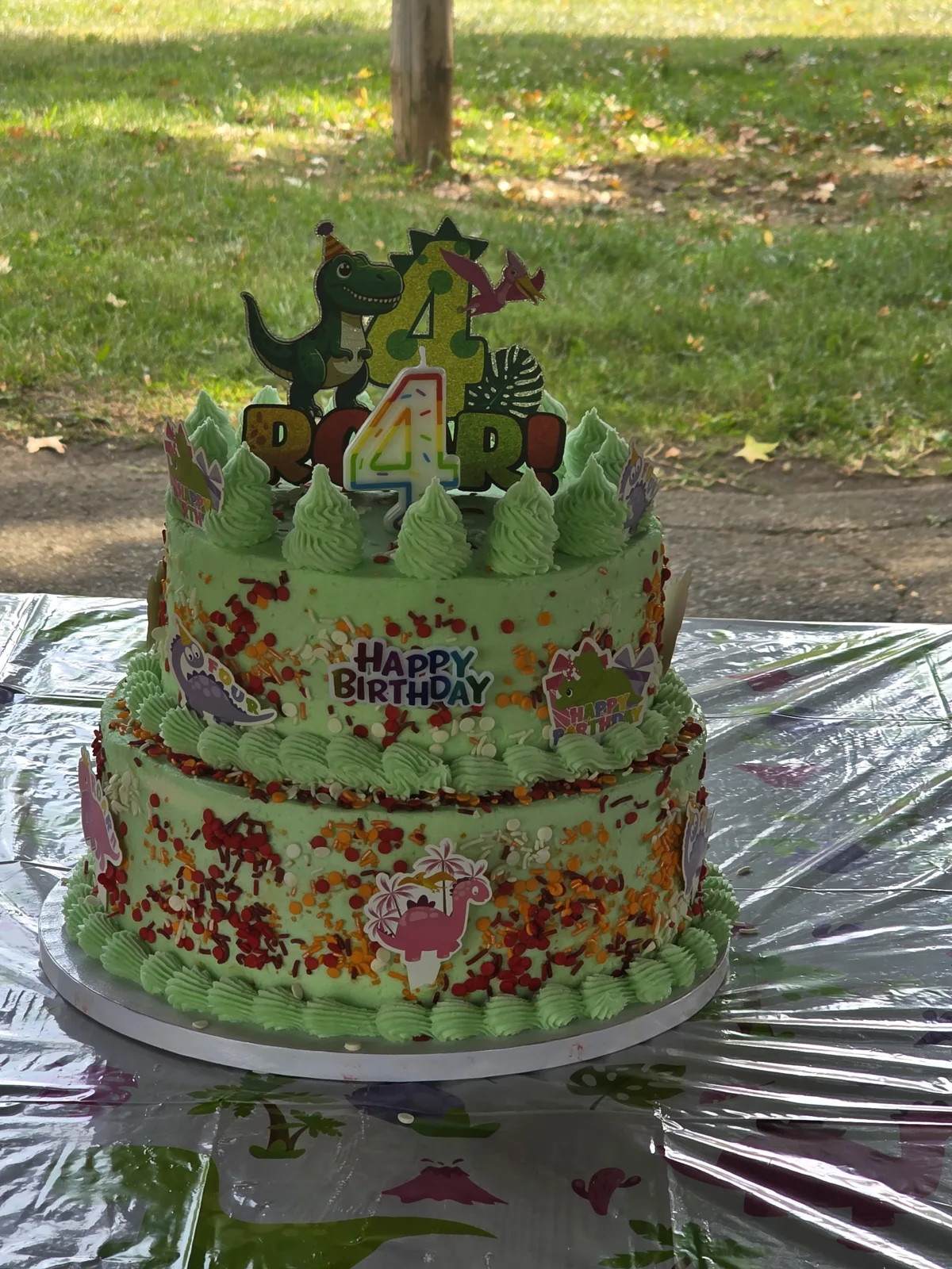Two tier green frosted birthday cake covered in red, yellow, and orange sprinkles with piped green borders and dinosaur themed decorations.