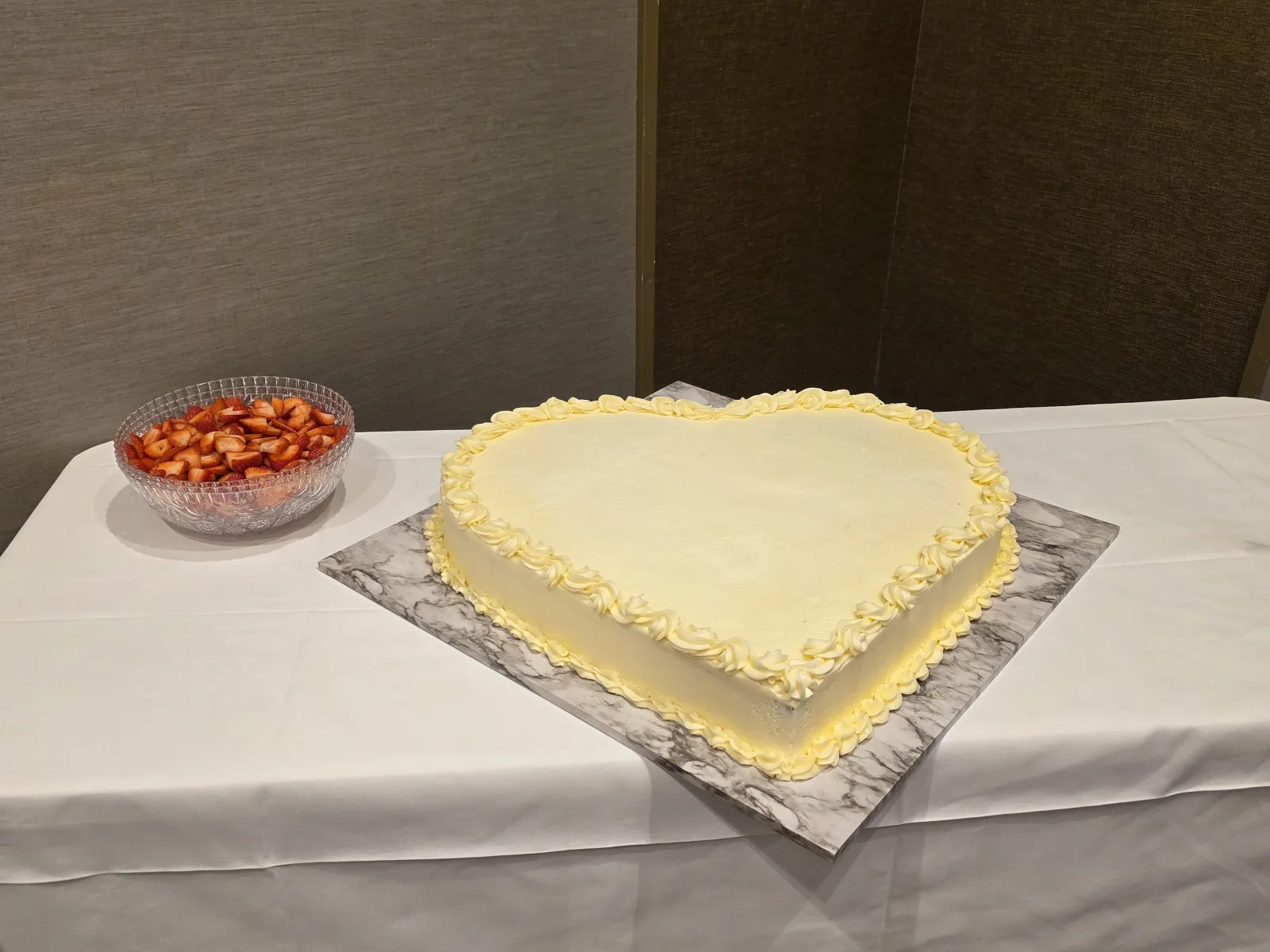 Large heart-shaped cake in pale yellow buttercream with a piped shell border on a marble board, beside a bowl of sliced strawberries.