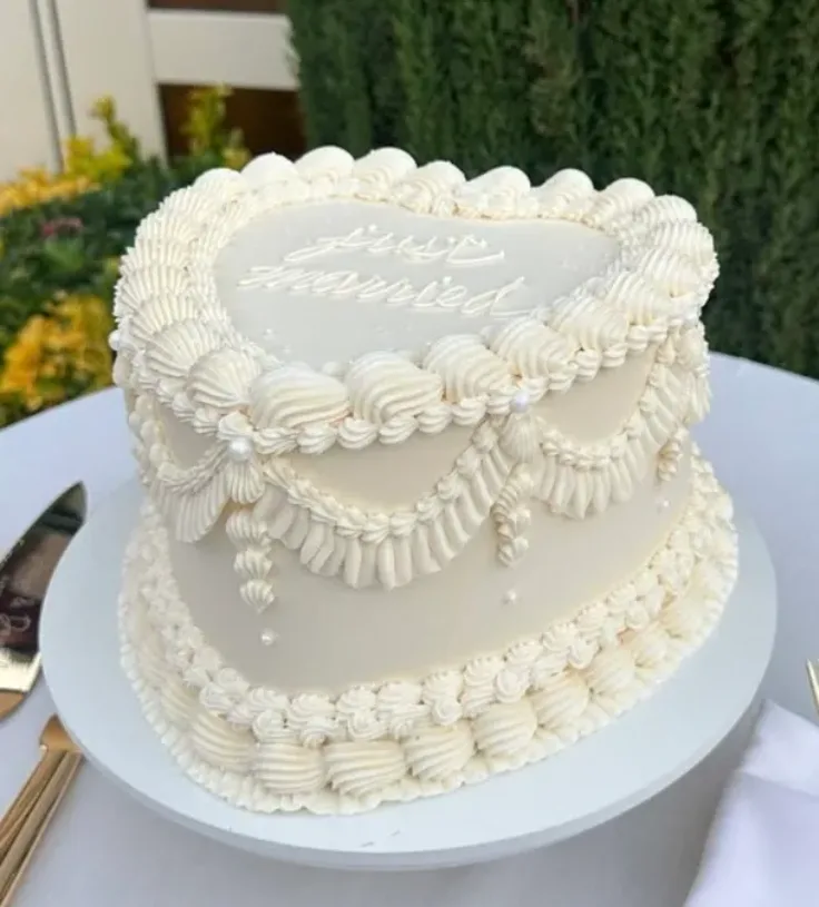 Two tiered white heart shaped wedding cake with piped buttercream swags, shell borders, pearl accents, and "Just Married" piped on top.