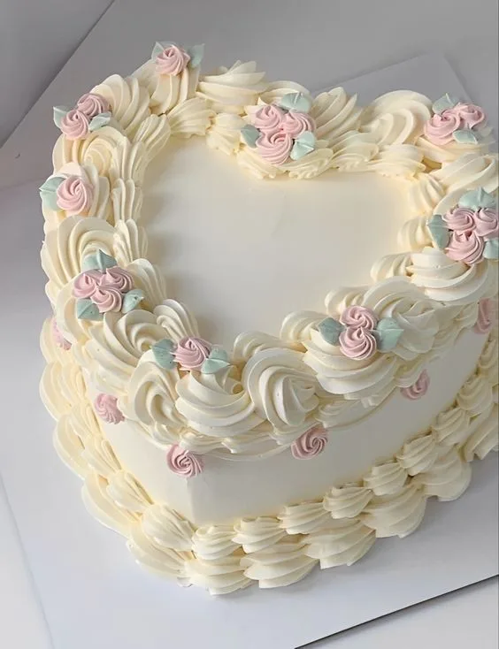 Heart shaped two tier cake covered in white buttercream with piped borders, pink rosette accents, and pale mint leaves around the edges.