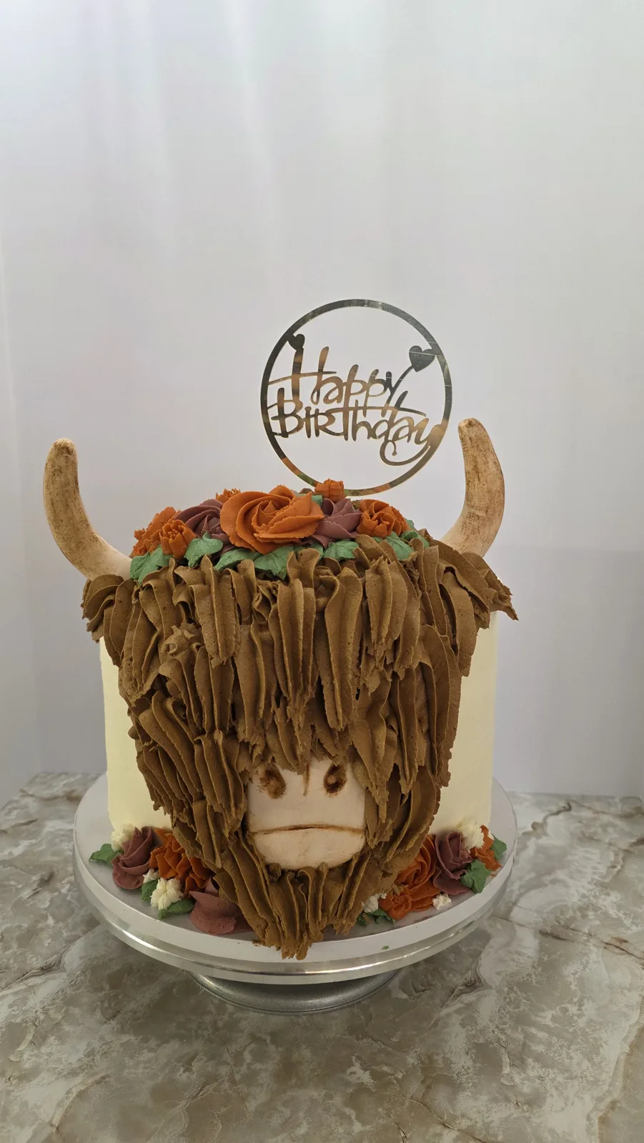 A round birthday cake decorated with brown textured frosting to resemble a shaggy Highland cow, featuring cream colored horns, a piped face, and orange and mauve buttercream roses on top and around the base, topped with a gold Happy Birthday topper.