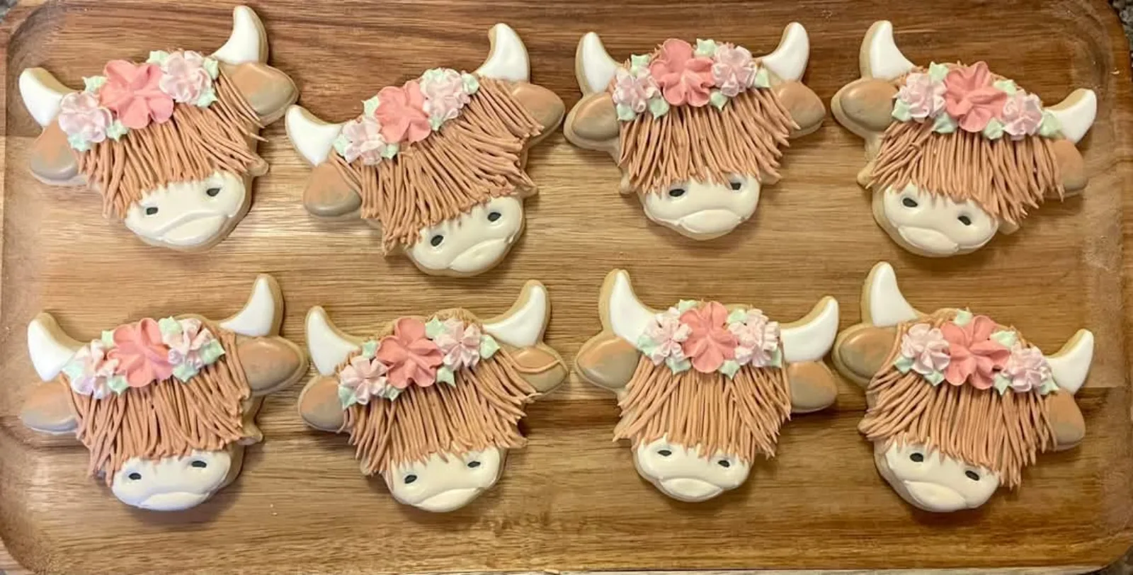 Eight highland cow cookies with piped fur and pink flower crowns on a wood board.
