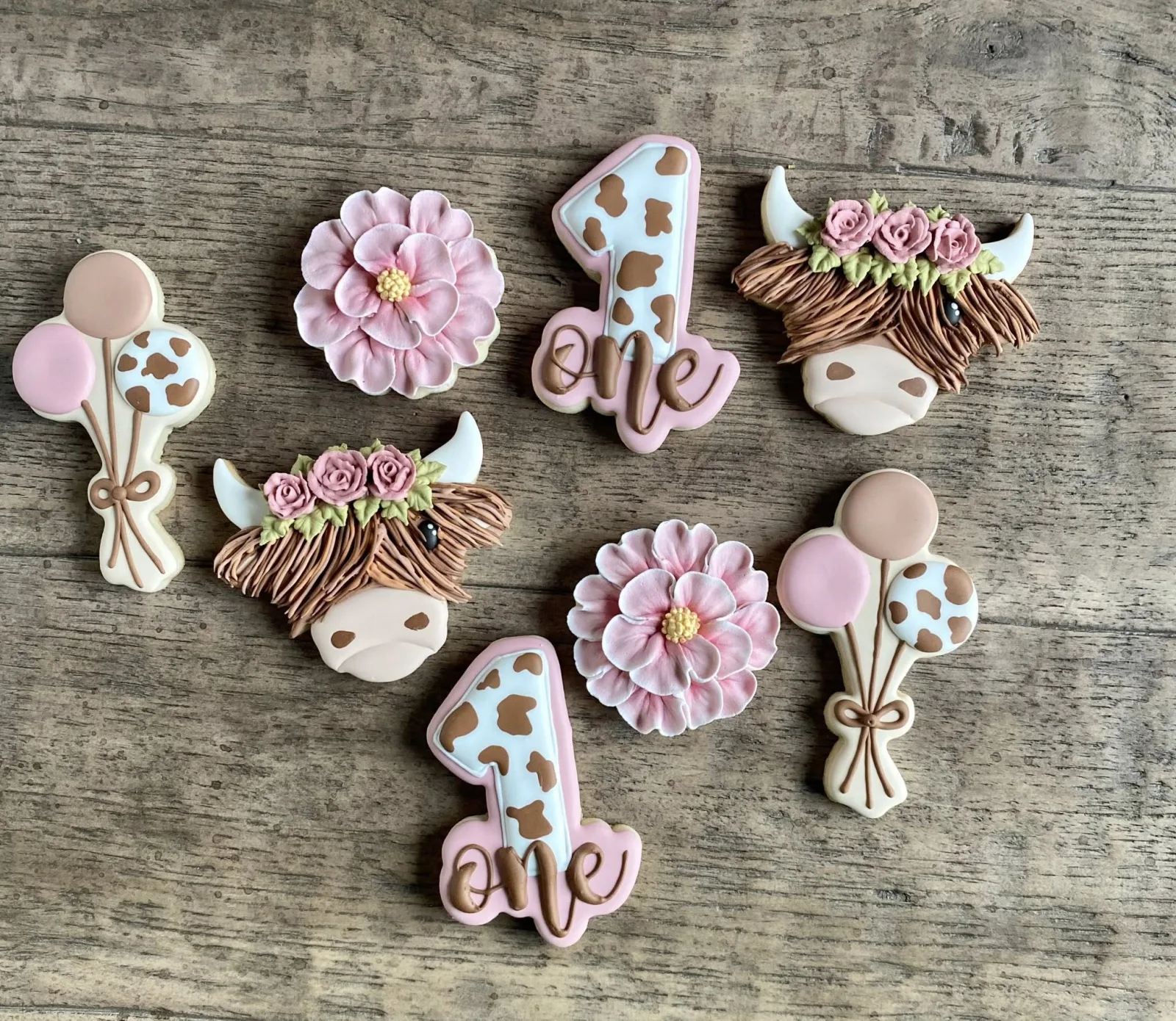First birthday cookies with highland cow faces, pink flowers, balloons and a number one, on weathered wood.