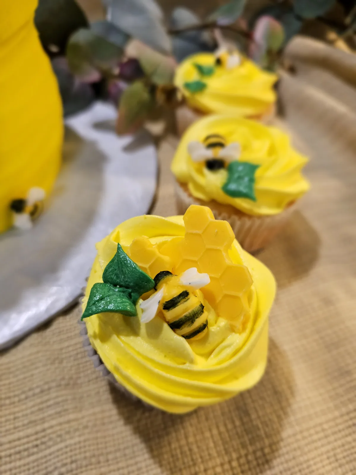Bright yellow swirled buttercream cupcakes topped with fondant honey bees, green leaves, and honeycomb accents arranged on a wooden surface.