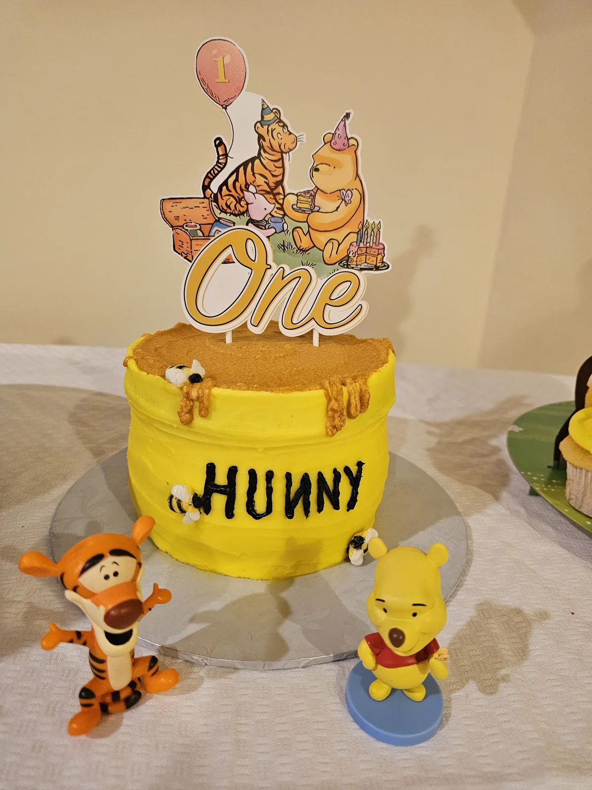 Yellow buttercream honey pot cake with brown honey drips, tiny bee decorations, and a Winnie the Pooh themed topper with a One balloon.