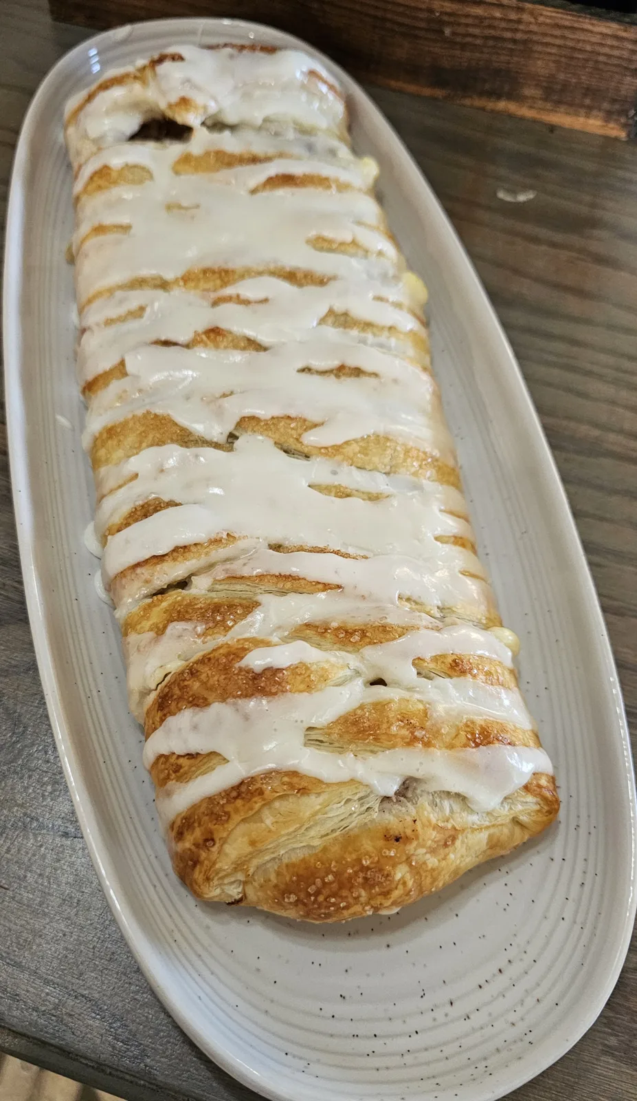 Golden brown braided pastry loaf with a flaky crust, drizzled with white icing, served on a white oval platter.