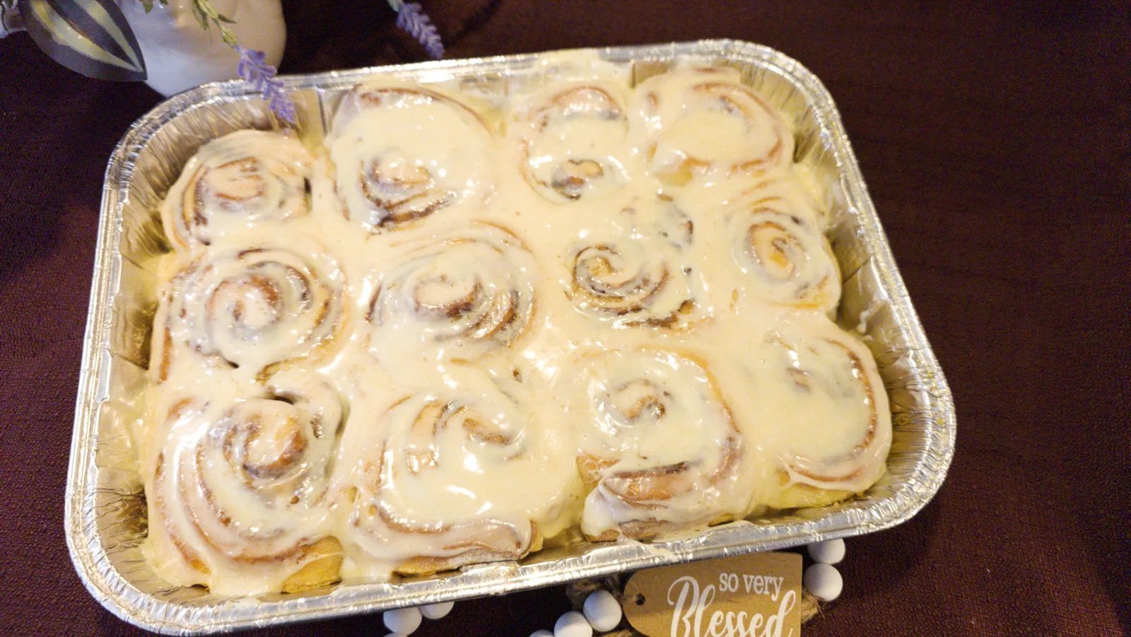 A dozen golden spiral cinnamon rolls in a square aluminum pan, generously topped with smooth white glaze icing.