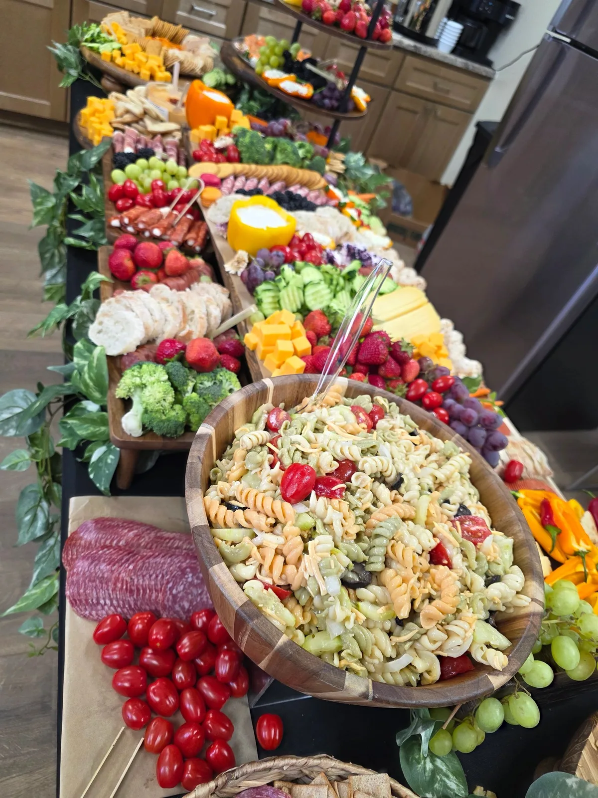 Elaborate grazing table featuring pasta salad, cubed cheese, salami, grapes, strawberries, broccoli, and bread on wooden boards.