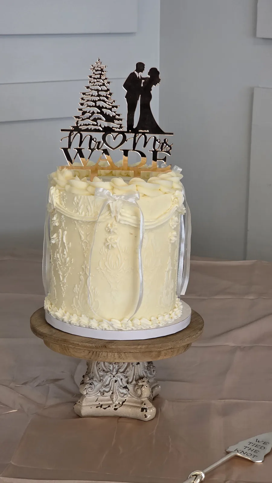 Single tier ivory wedding cake featuring an embossed lace pattern, draped fondant bow detail, and a black silhouette couple and tree cake topper.