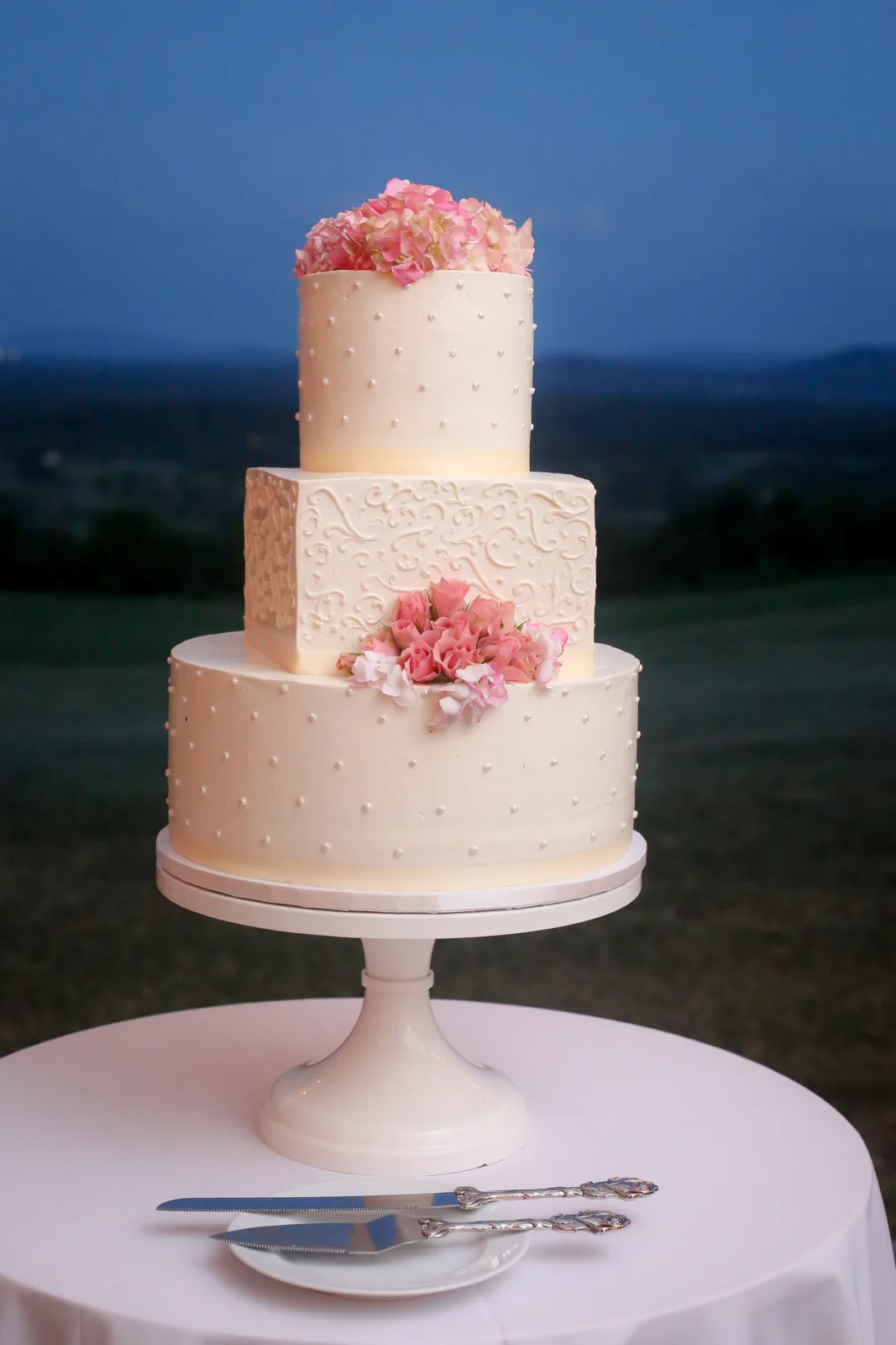 Three tier ivory wedding cake with round top and bottom tiers dotted with pearls, a square middle tier with embossed scrollwork, topped with pink and peach floral arrangements.