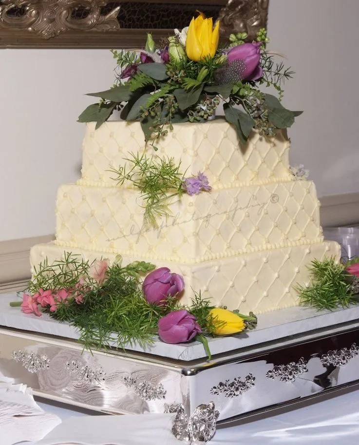 Three tier ivory wedding cake with quilted lattice pattern, pearl beading, and fresh purple, pink, and yellow floral topper with greenery on silver stand.