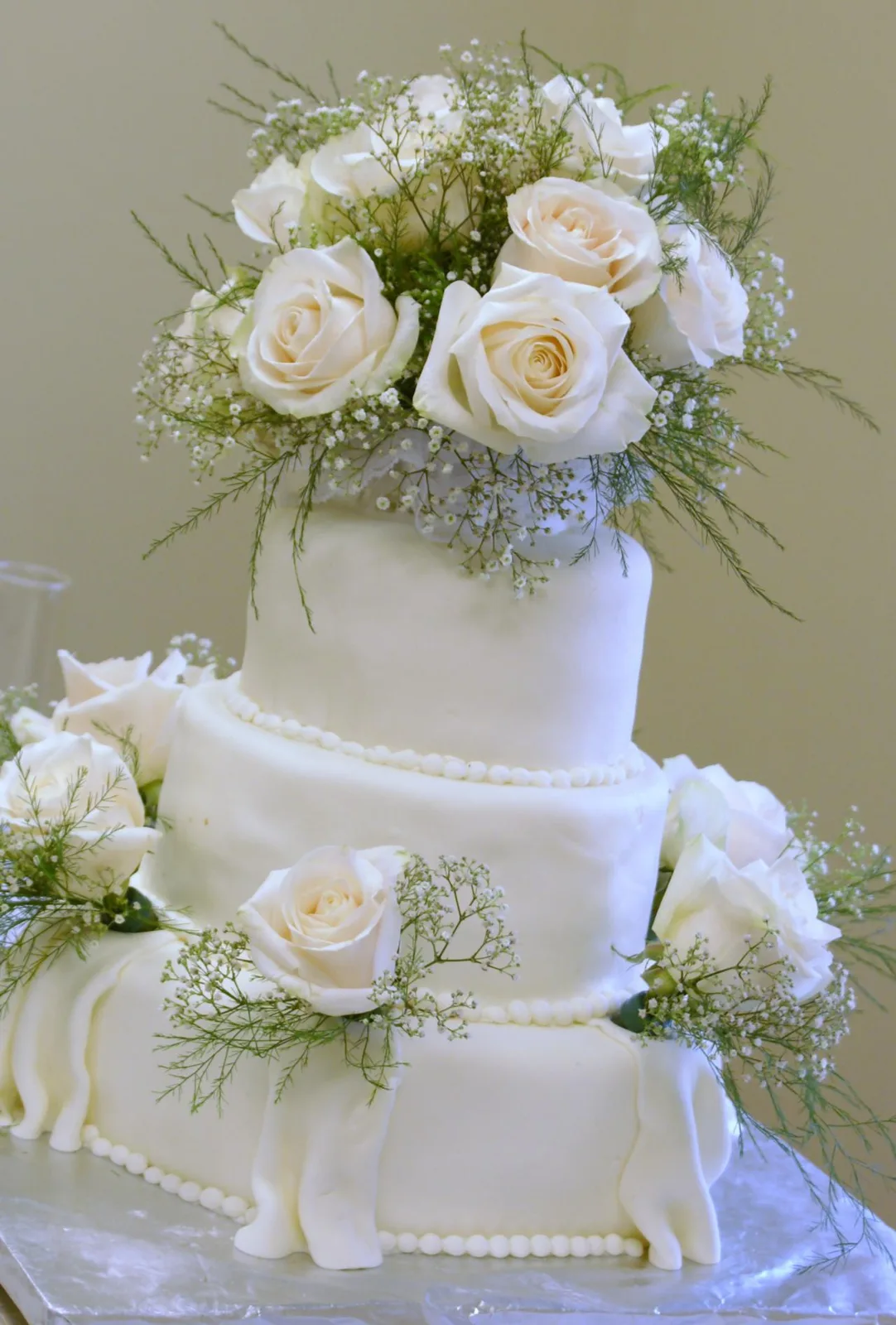 Three tiered white wedding cake with draped fondant details, beaded borders, and topped with cream colored roses and baby's breath.