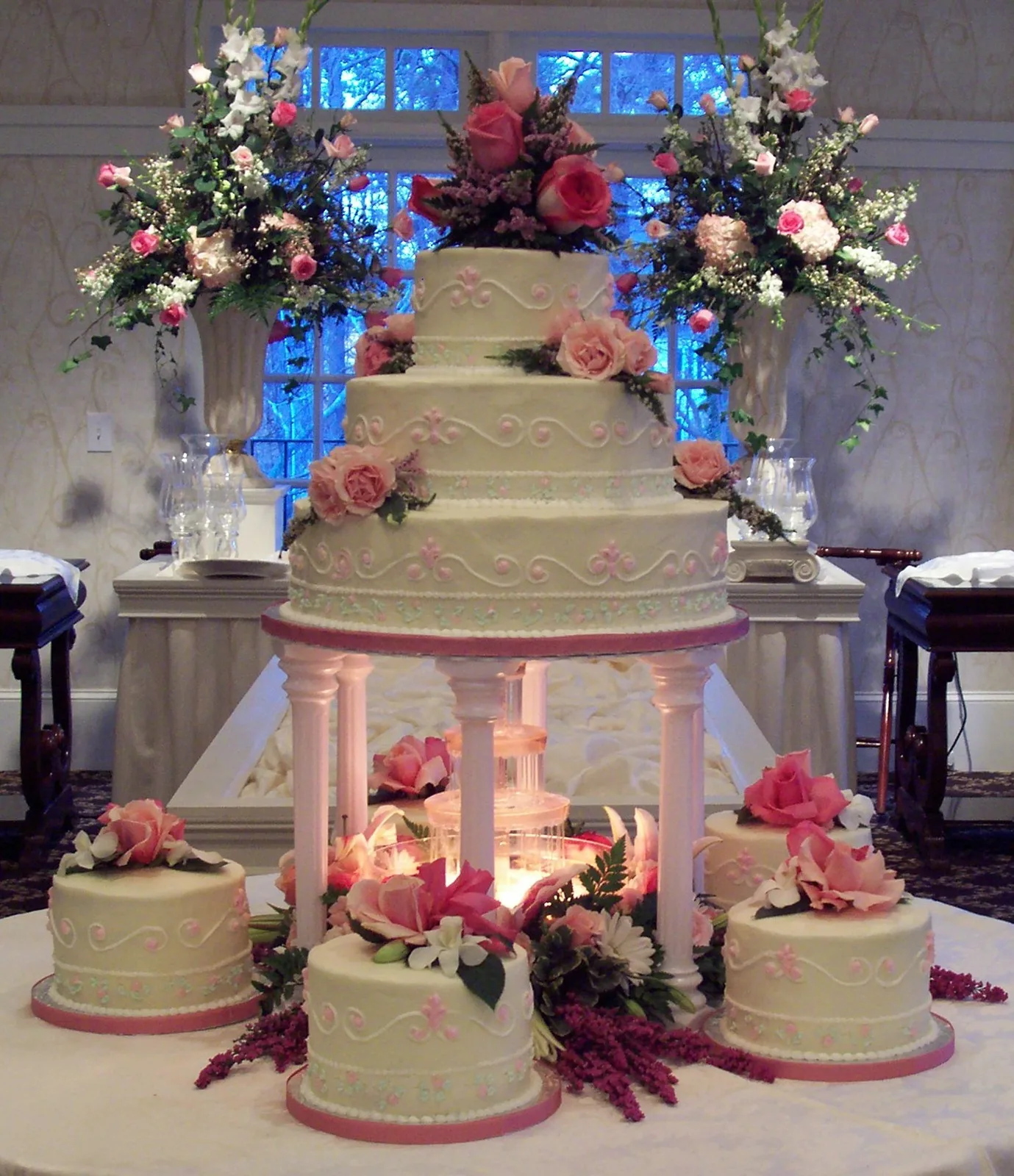 Four tier ivory buttercream cake with pink scroll piping and pink rose clusters, displayed on columns surrounded by matching smaller cakes.