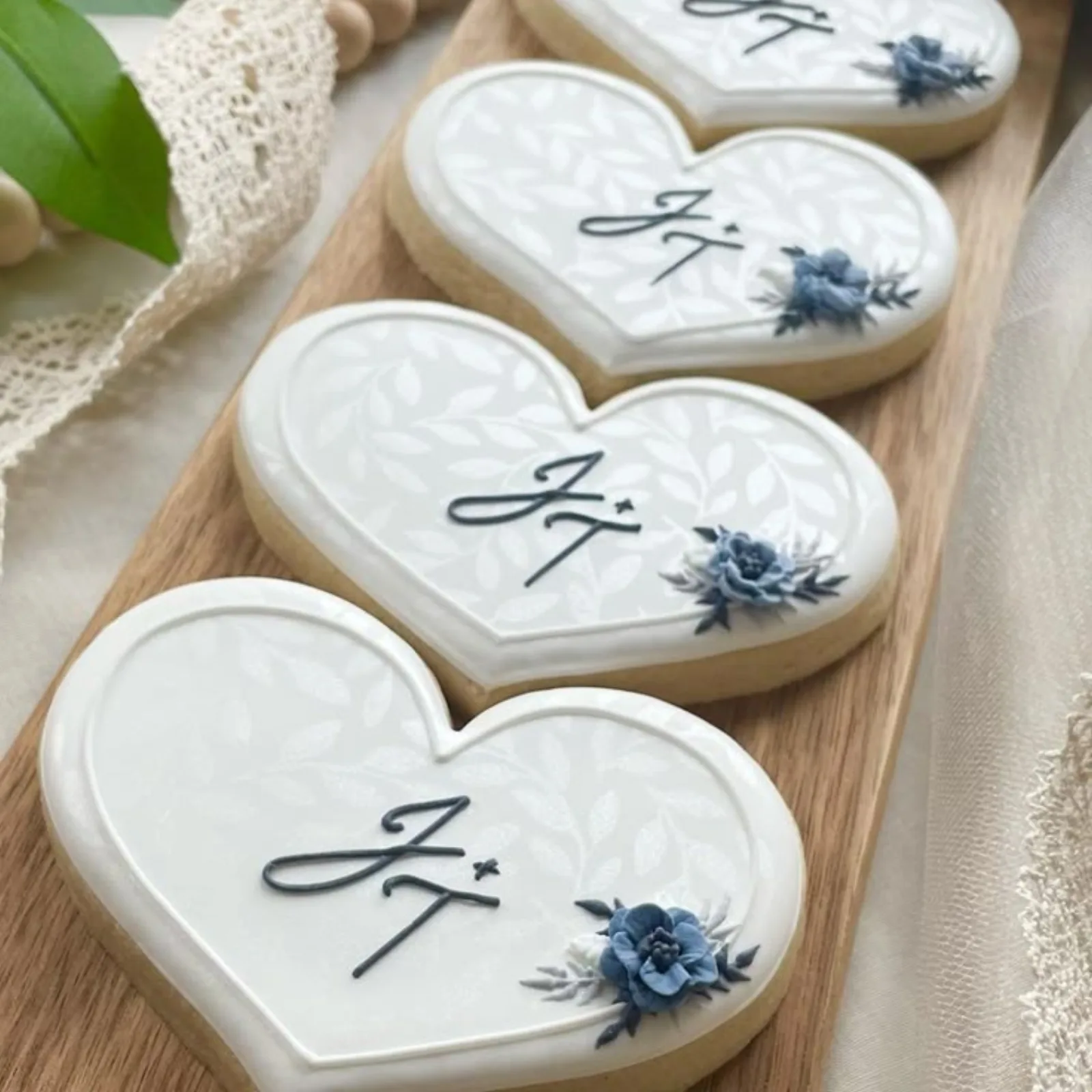 White heart cookies with a JT monogram, a leaf pattern and blue flowers on a wood board.