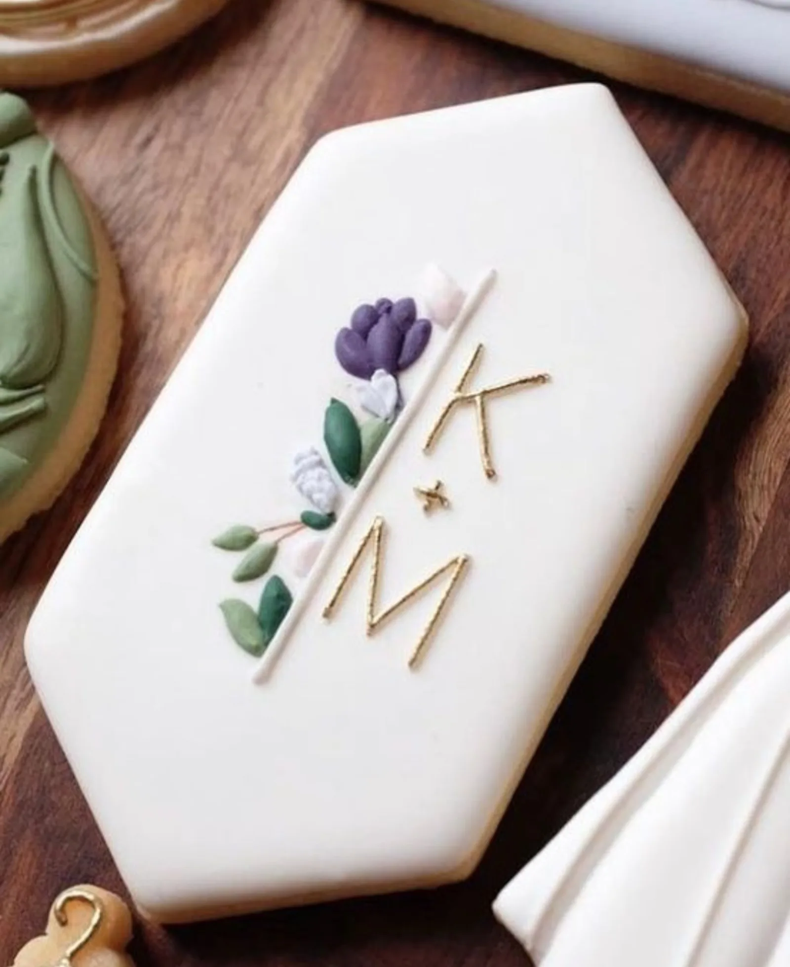 White hexagon cookie with a gold K and M monogram, a purple tulip and green leaves on wood.