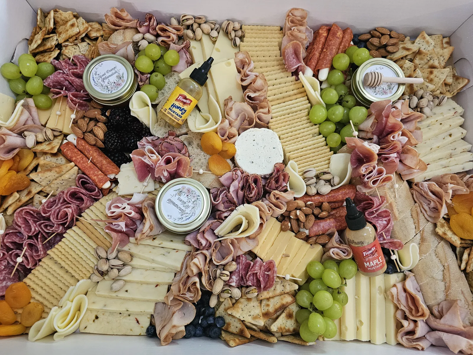 A packed white charcuterie box filled with sliced cheeses, rolled cured meats, green and black grapes, assorted crackers, almonds, pistachios, dried apricots, blueberries, jam jars, and mustard bottles.