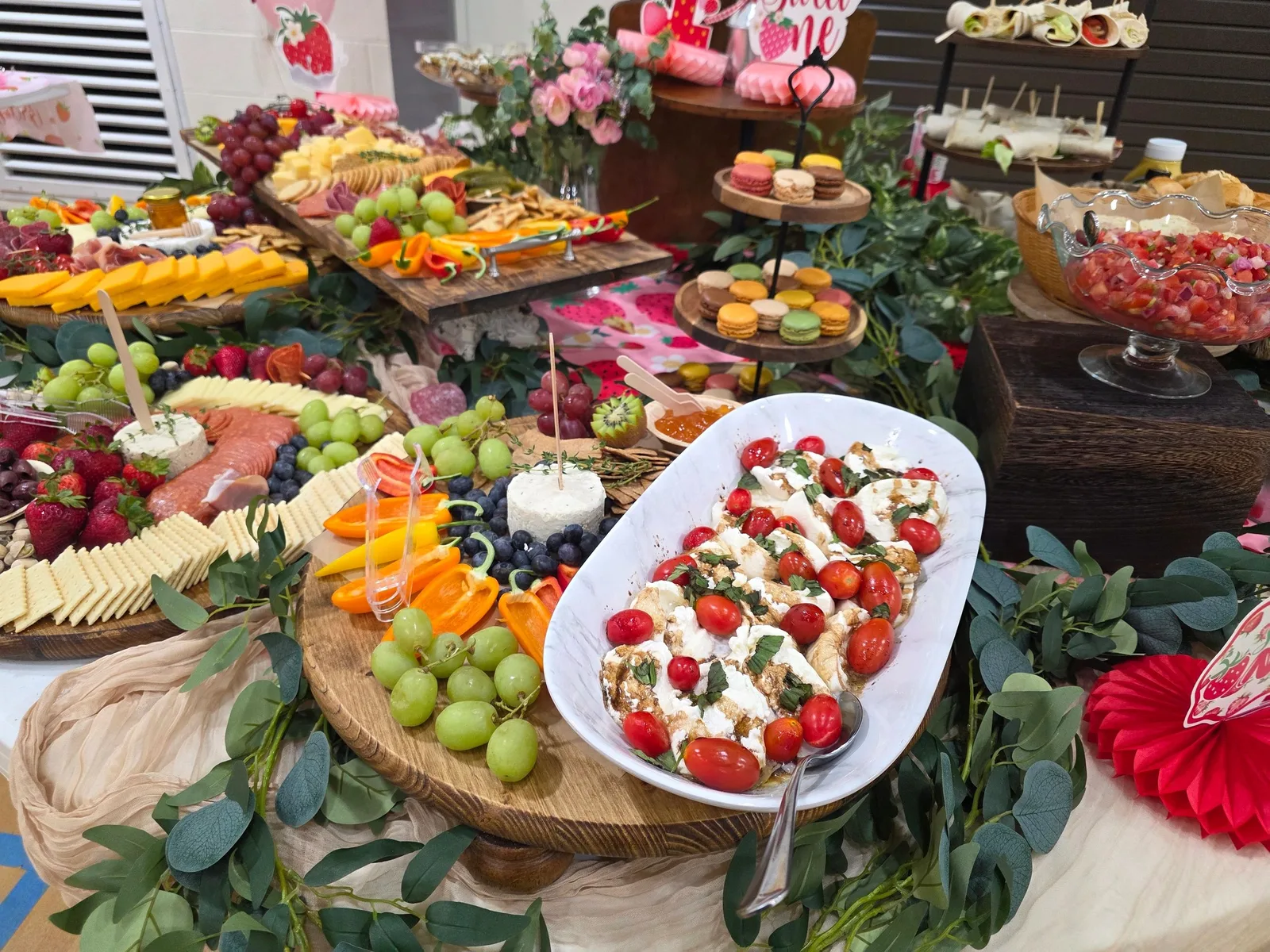 A lavish party spread featuring charcuterie boards with meats, cheeses, and fruits, a tomato-topped salad, macarons, pinwheel wraps, and pink cookies.