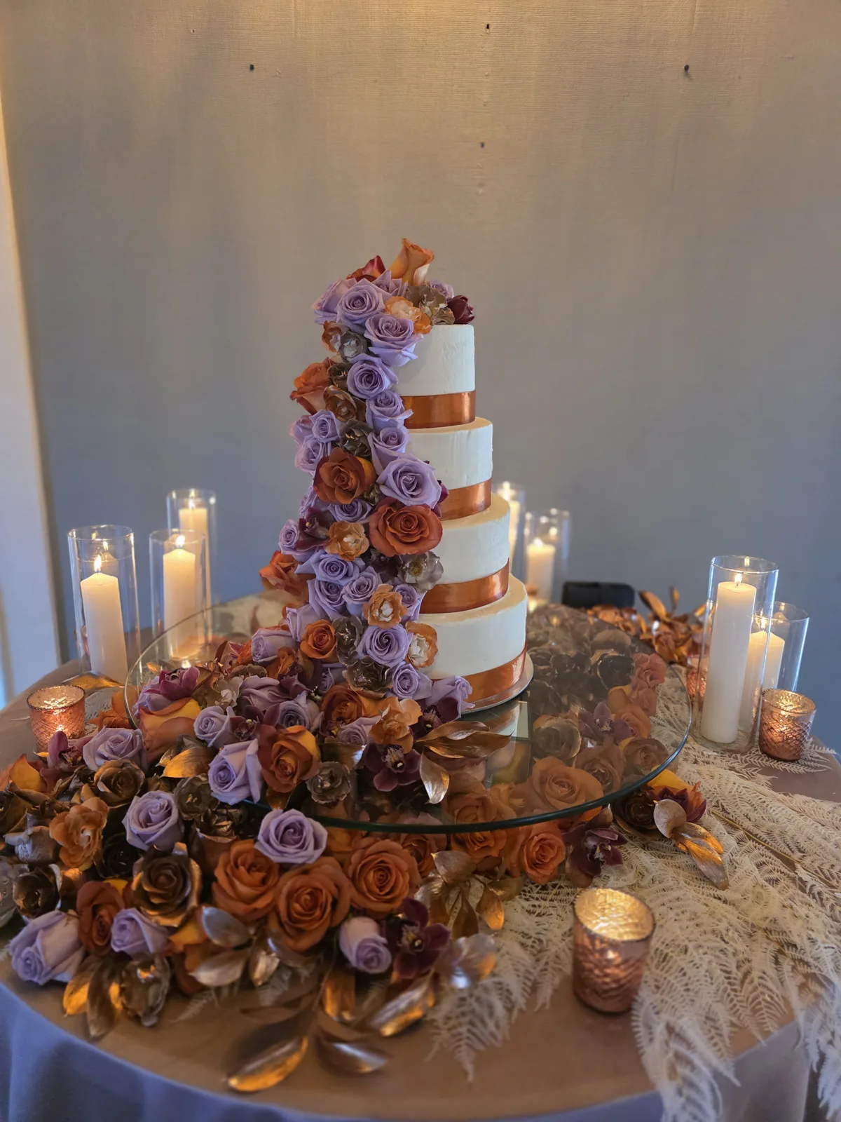 A five tier white wedding cake with copper ribbon trim on each tier, decorated with a cascading arrangement of lavender purple, rust orange, and burgundy roses down one side.