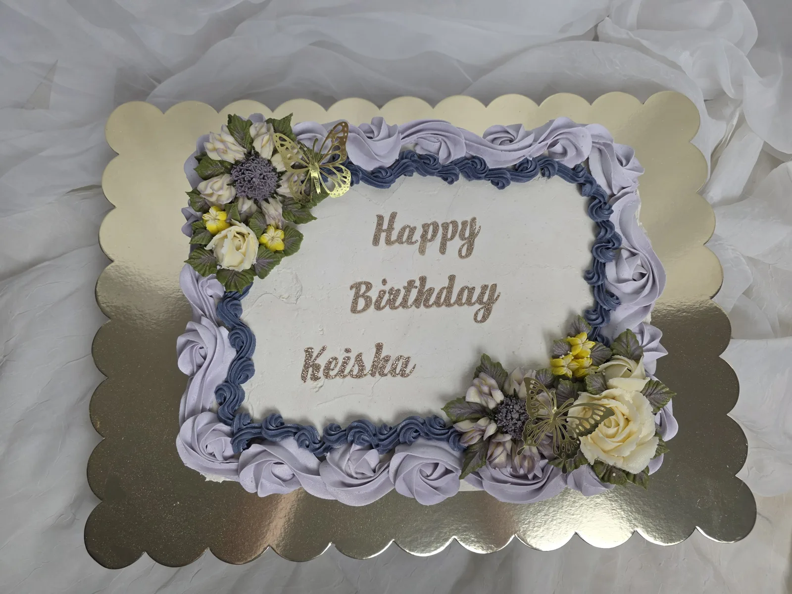 Rectangular white frosted sheet cake with lavender and navy blue piped rose border, cream and yellow floral clusters, and gold butterfly accents on a scalloped gold board.