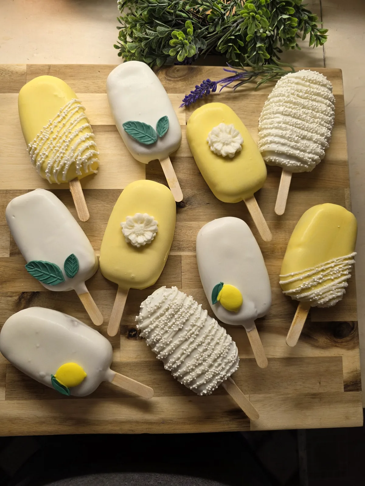 Yellow and white cakesicles decorated with fondant flowers, lemon slices, green leaves, and white sprinkle drizzle arranged on a wooden board.