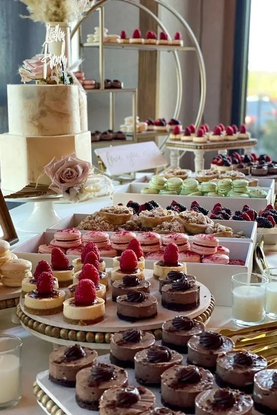 Dessert table with a tiered cake, macarons, raspberry tarts, and chocolate mousse cups at a wedding reception.