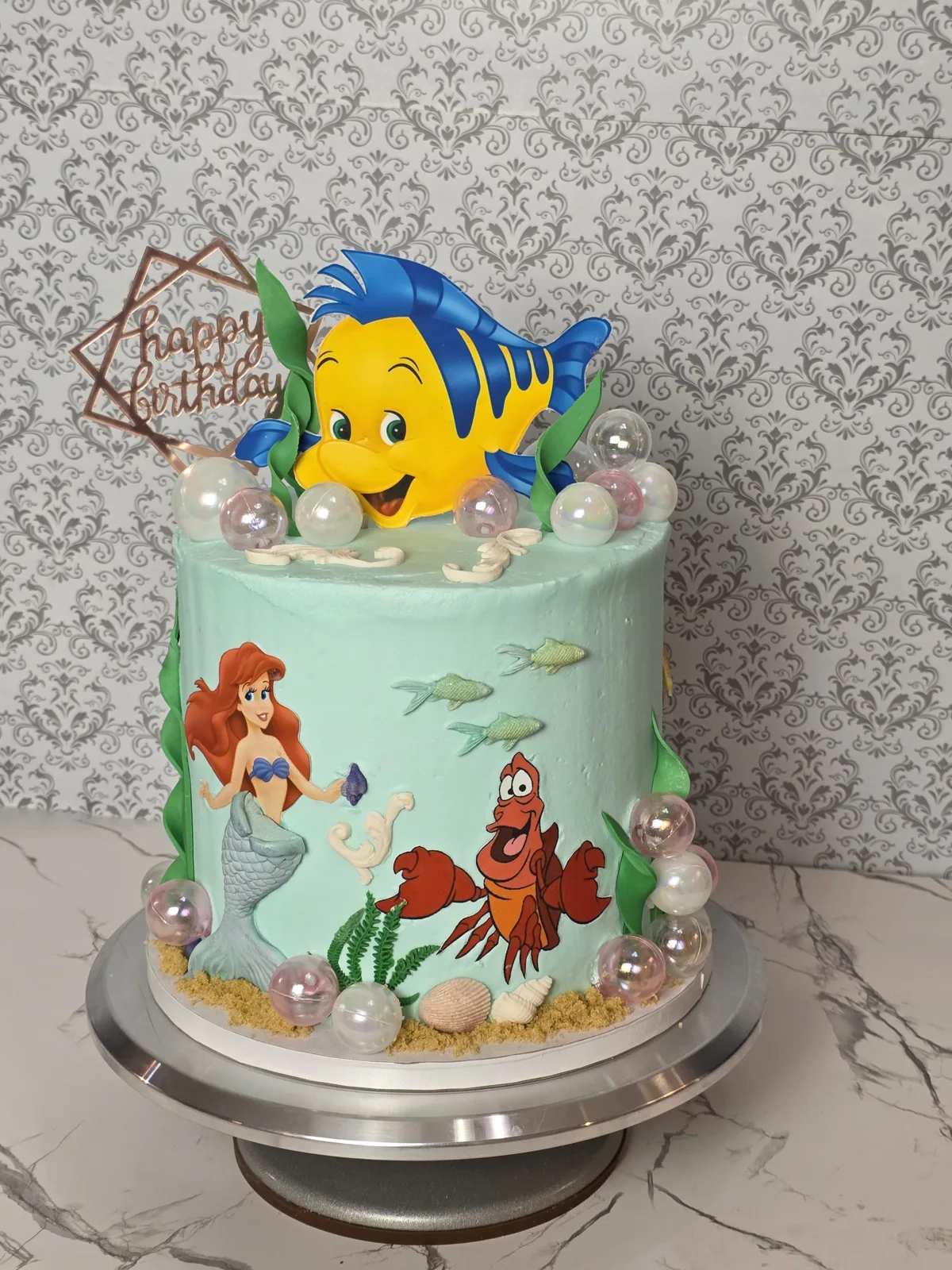 A seafoam green cake features mermaid and crab designs, a large yellow and blue fish topper, clear bubble spheres, and a Happy Birthday sign.