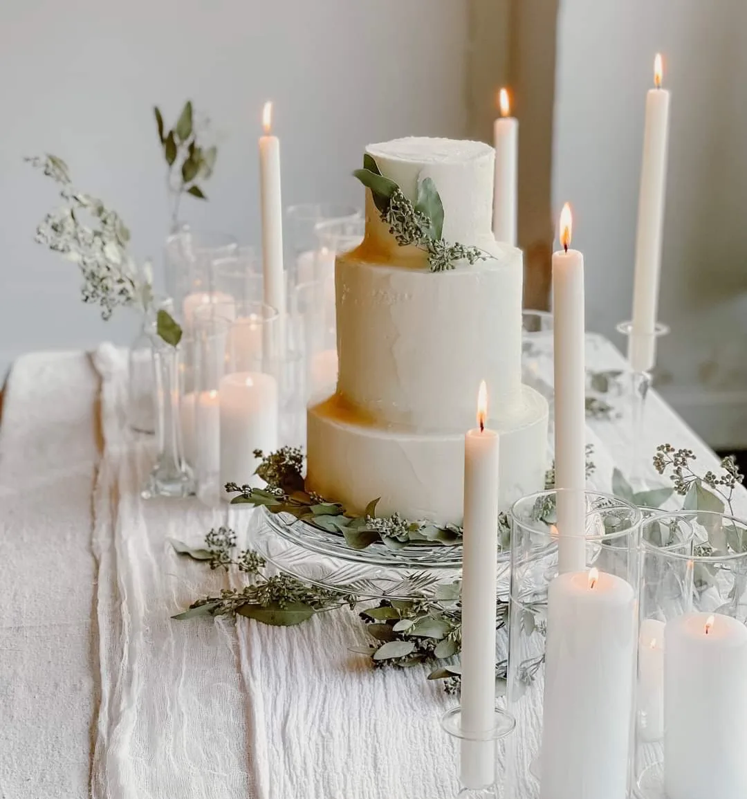 Three tier round white cake with textured buttercream and fresh eucalyptus sprigs displayed on a crystal stand surrounded by lit taper candles.