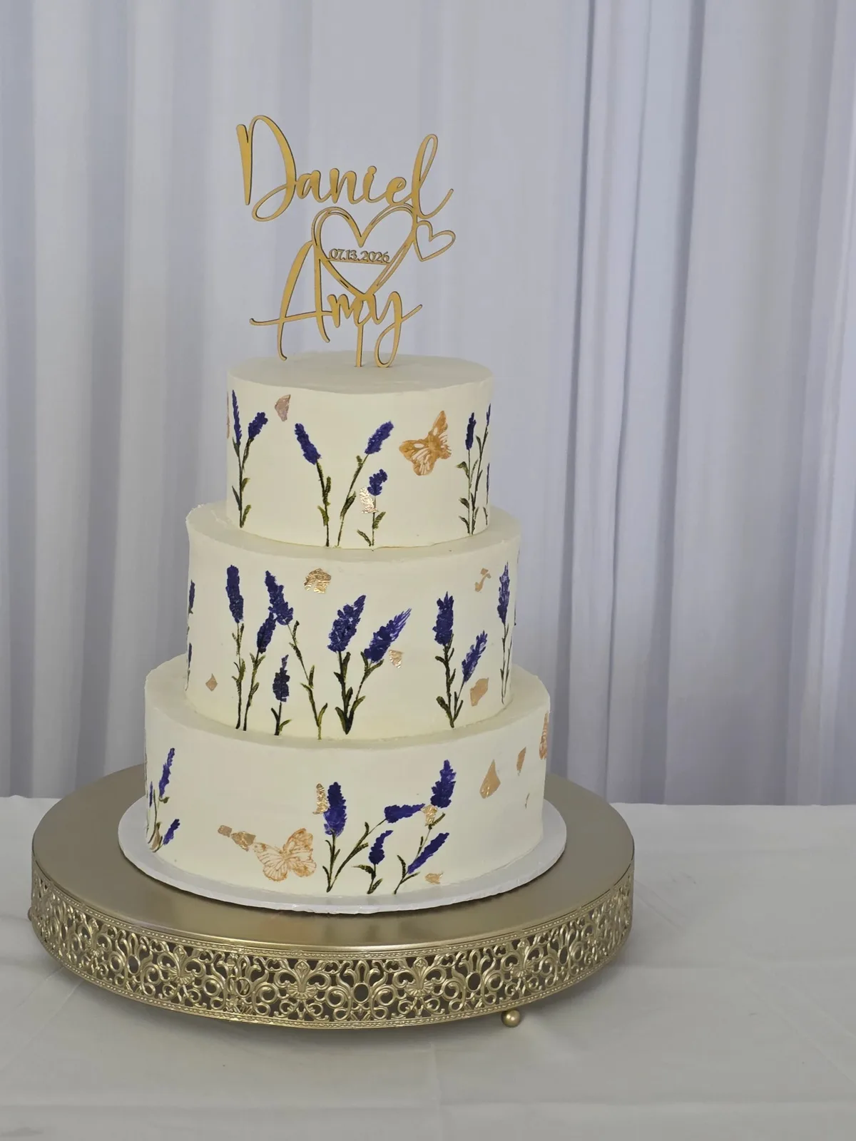 Three-tiered white cake decorated with painted purple flowers, green stems, and gold butterflies.