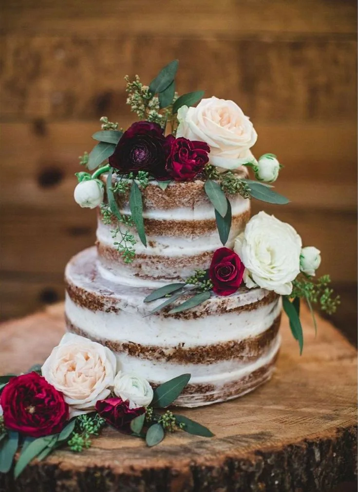 A two tier naked cake with thin white frosting over brown sponge, adorned with cream roses, burgundy blooms, and eucalyptus on a wooden slice.