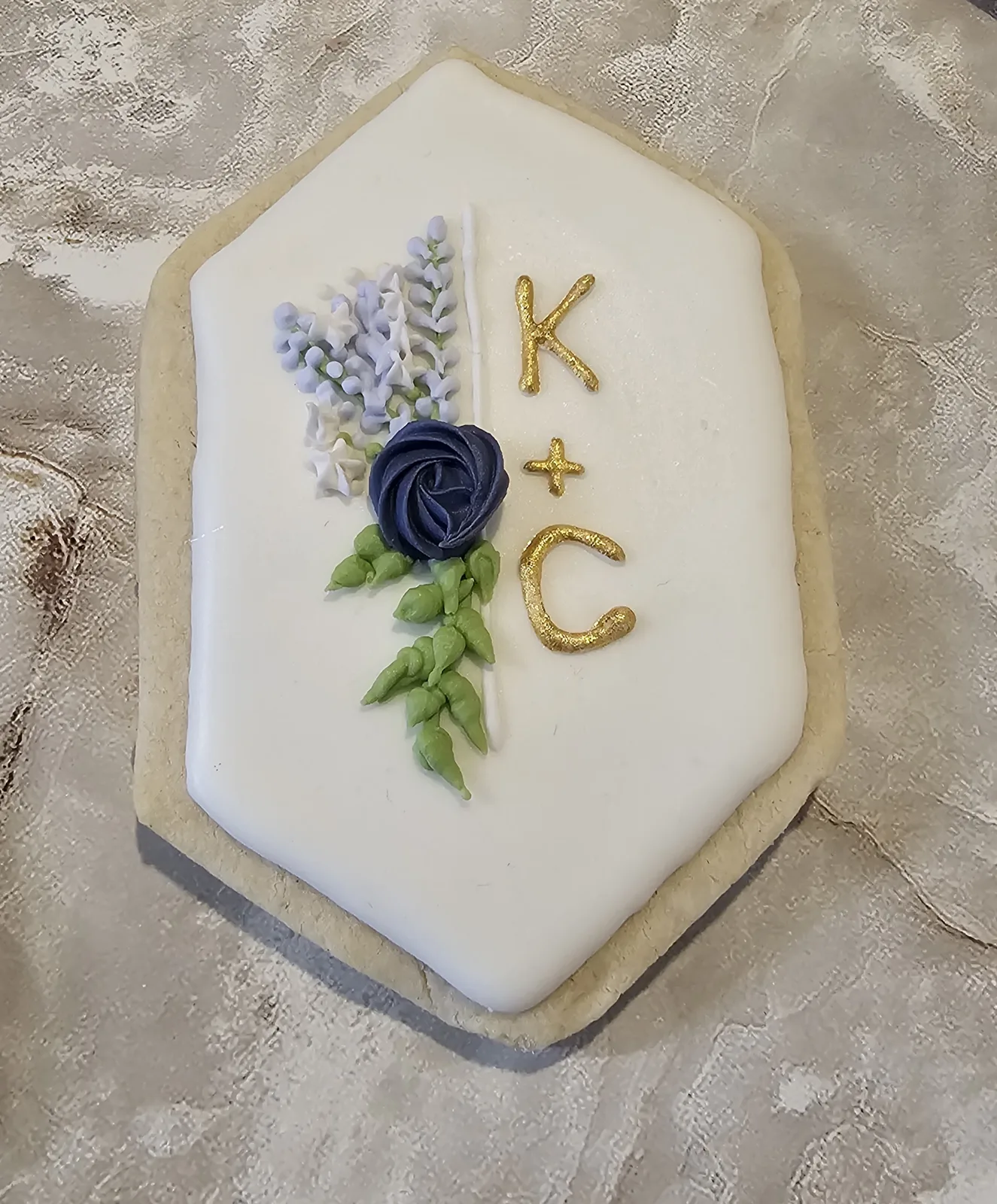 White iced sugar cookie decorated with piped lavender florets, a navy rose with green leaves, and metallic gold initials K and C separated by a small cross.