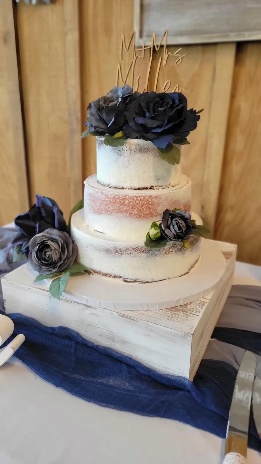 Three tier semi naked white wedding cake with copper glitter accents, navy blue and gray roses, and a wooden Mr and Mrs topper on a white wooden crate.