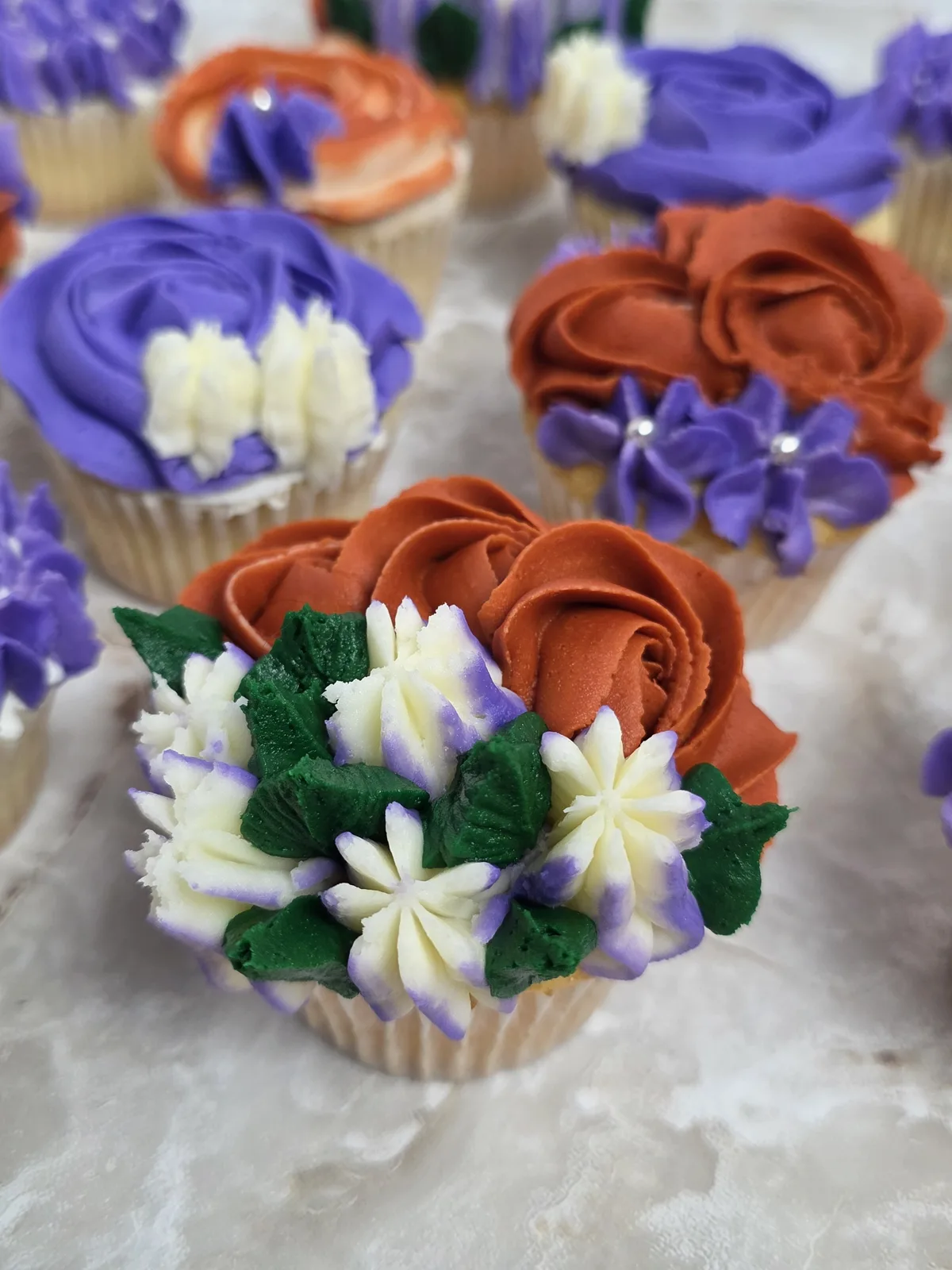 Cupcake with piped orange rose frosting, green leaves, and purple tipped white flowers, surrounded by other purple and orange frosted cupcakes.